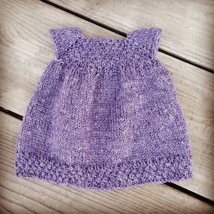 Puede incluir: Un vestido de bebé de punto de color morado con un diseño sencillo y un dobladillo festoneado.