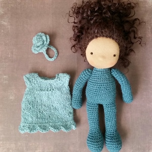 May include: A handmade doll with a teal knitted body and brown curly hair. The doll has a blank face and is accompanied by a teal knitted dress and a flower-shaped hair accessory. The doll is approximately 12 inches tall.