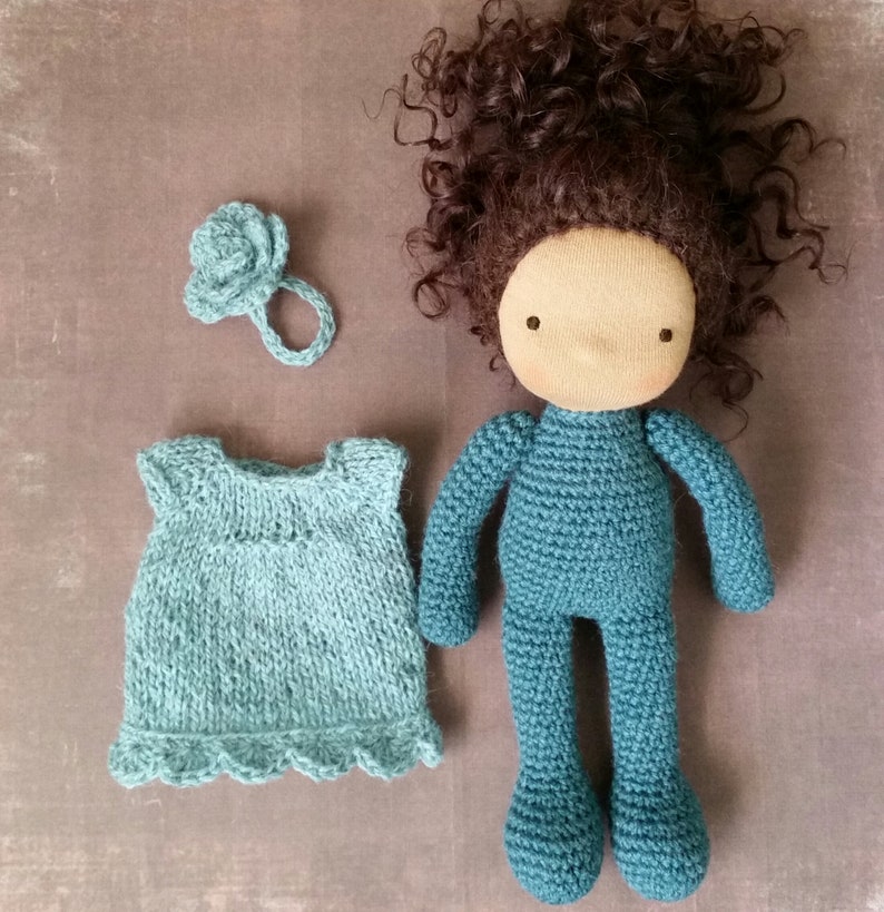 May include: A handmade doll with a teal knitted body and brown curly hair. The doll has a blank face and is accompanied by a teal knitted dress and a flower-shaped hair accessory. The doll is approximately 12 inches tall.