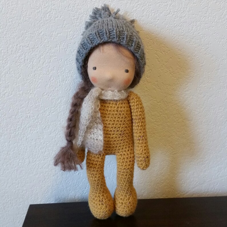 May include: A handmade doll with a mustard yellow crocheted body, wearing a gray knit hat and a white and gray scarf. The doll has a fabric face with rosy cheeks and a long brown braid.
