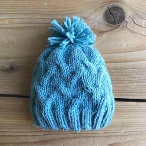 Puede incluir: Un gorro azul de punto con un patrón de trenza y un pompón esponjoso en la parte superior.