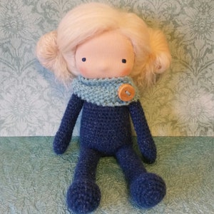May include: Handmade doll with a fabric face, light blonde yarn hair, and a blue knitted outfit. The doll has a light blue knitted scarf with a wooden button closure. The doll's body and limbs are made of a dark blue knitted fabric.