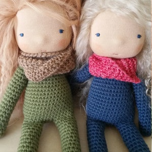 May include: Two handmade cloth dolls with long, flowing hair. One doll is dressed in green crochet, the other in blue. Both dolls have tan faces, blue eyes, and knitted scarves. The dolls are approximately 12 inches tall.