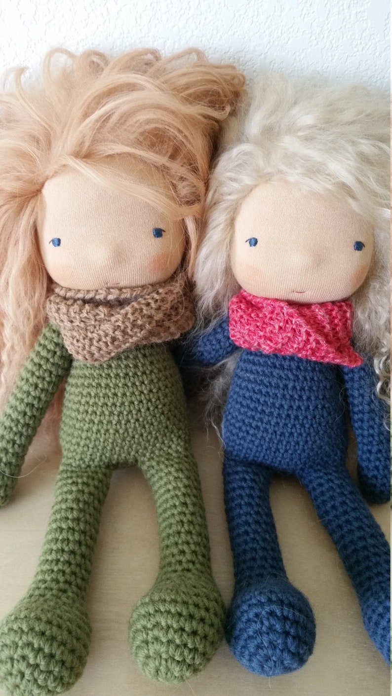 May include: Two handmade cloth dolls with long, flowing hair. One doll is dressed in green crochet, the other in blue. Both dolls have tan faces, blue eyes, and knitted scarves. The dolls are approximately 12 inches tall.
