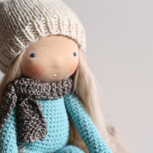 May include: A handmade doll with a light skin tone, blue eyes, and long blonde hair. The doll wears a cream-colored knitted hat, a gray scarf, and a turquoise blue knitted sweater. The doll is a soft toy.