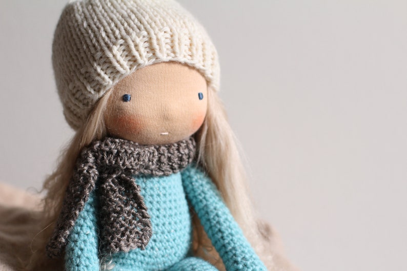 May include: A handmade doll with a light skin tone, blue eyes, and long blonde hair. The doll wears a cream-colored knitted hat, a gray scarf, and a turquoise blue knitted sweater. The doll is a soft toy.