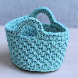 Puede incluir: Una pequeña cesta de crochet azul claro con dos asas. La cesta está hecha con un punto texturizado y tiene una forma redondeada.