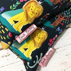 May include: Close-up of two fabric items with a dark background featuring a whimsical lion design in yellow and a floral and tree pattern. Pink labels with the text "Book Buddy" are visible. The items appear to be handmade.