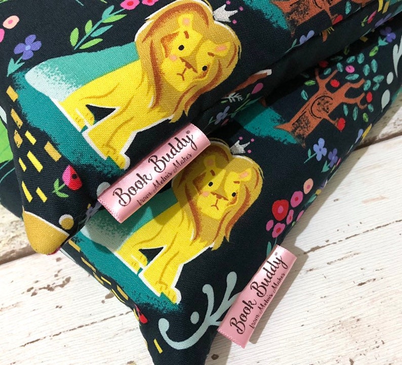 May include: Close-up of two fabric items with a dark background featuring a whimsical lion design in yellow and a floral and tree pattern. Pink labels with the text "Book Buddy" are visible. The items appear to be handmade.