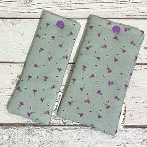 May include: Two rectangular fabric cases with a light green background, adorned with a pattern of purple thistles and small white dots. Each case has a purple button closure. The label "Melvis Maker" is visible on each case.