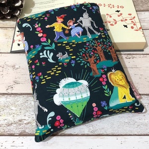 May include: A dark blue book sleeve featuring a colorful illustration of characters from The Wizard of Oz. The design includes Dorothy, the Scarecrow, the Tin Man, the Cowardly Lion, and a hot air balloon, surrounded by flowers and trees.
