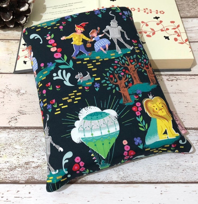 May include: A dark blue book sleeve featuring a colorful illustration of characters from The Wizard of Oz. The design includes Dorothy, the Scarecrow, the Tin Man, the Cowardly Lion, and a hot air balloon, surrounded by flowers and trees.