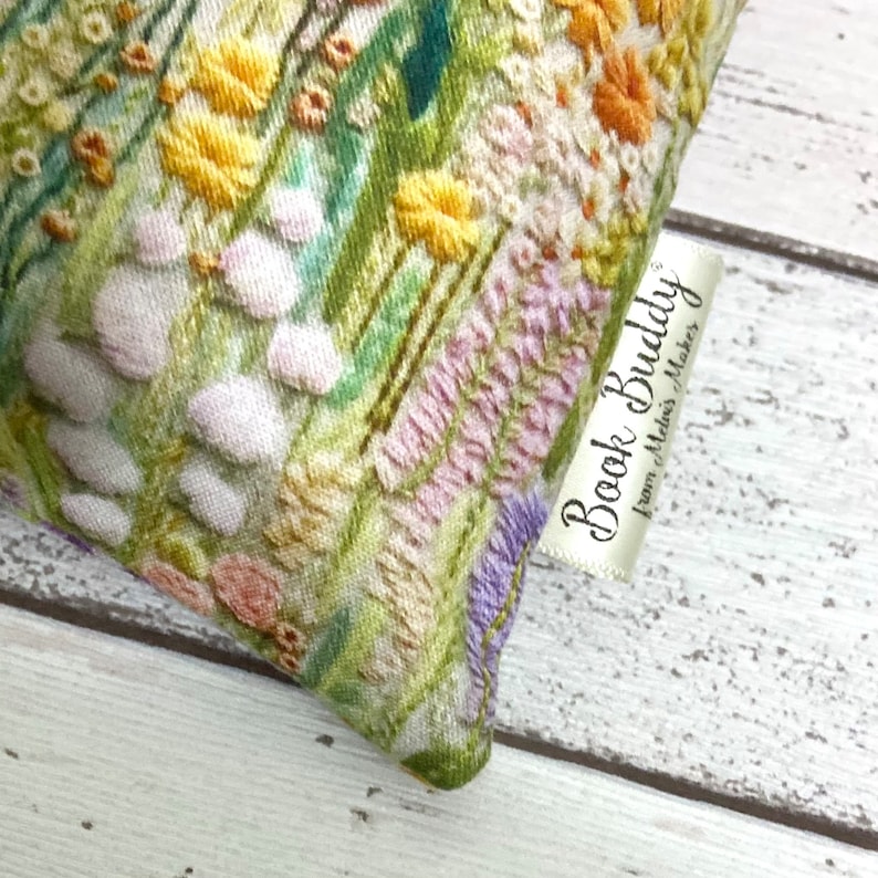 Puede incluir: Una bolsa de tela con estampado floral verde, blanco y morado con una etiqueta blanca que dice "Book Buddy from Stitches Makers".