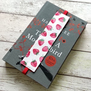 May include: A white bookmark with red strawberries printed on it. The bookmark has a red ribbon attached to it. The bookmark is on top of a gray book with the title "To Kill a Mockingbird" printed on the cover.