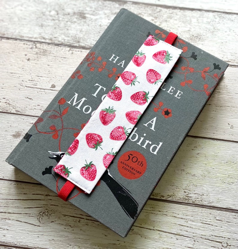May include: A white bookmark with red strawberries printed on it. The bookmark has a red ribbon attached to it. The bookmark is on top of a gray book with the title "To Kill a Mockingbird" printed on the cover.