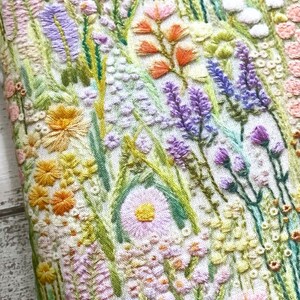 Puede incluir: Un primer plano de una tela blanca con flores bordadas en tonos de rosa, morado, amarillo y verde. Las flores est&aacute;n dispuestas en un patr&oacute;n aleatorio y crean un dise&ntilde;o hermoso y delicado.