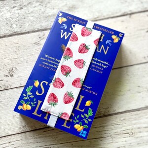 May include: A white bookmark with a red strawberry pattern. The bookmark is attached to a blue book with the title "Still Life" and the text "The Sunday Times Bestseller".
