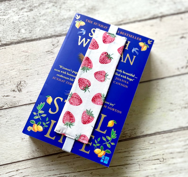 May include: A white bookmark with a red strawberry pattern. The bookmark is attached to a blue book with the title "Still Life" and the text "The Sunday Times Bestseller".