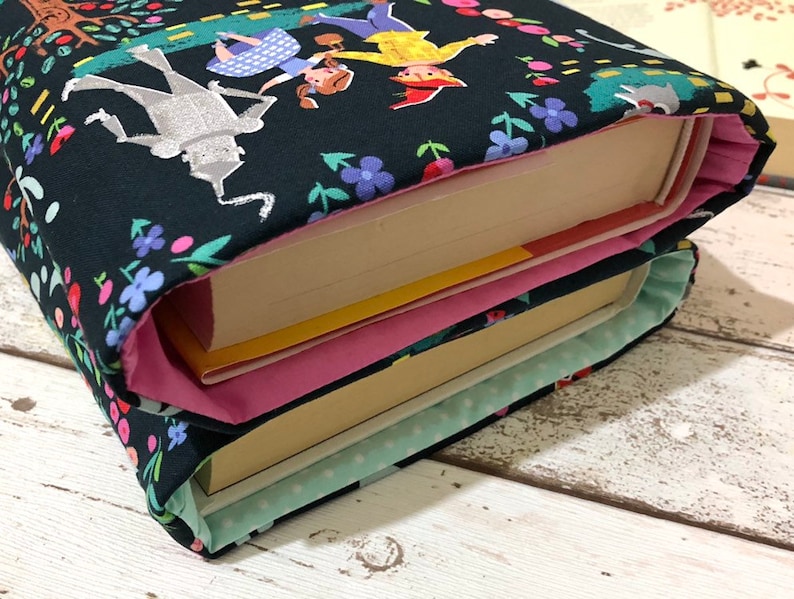 May include: Two book covers with a dark background featuring a colorful illustration of characters from The Wizard of Oz. The covers are decorated with floral patterns and have pink and light blue linings.