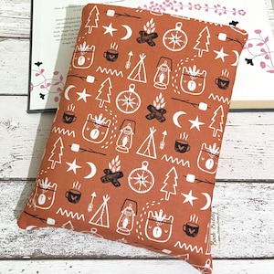 May include: An orange fabric book sleeve featuring a white camping design. The pattern includes trees, bears, campfires, lanterns, compasses, and stars. The sleeve is on a wooden surface with a book.
