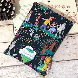 May include: A dark blue book sleeve with a vibrant illustration of characters from The Wizard of Oz. The design includes Dorothy, the Tin Man, the Scarecrow, the Cowardly Lion, and Toto, set against a floral and tree-filled background.