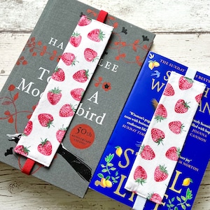 May include: Two book marks with a white background and red strawberries printed on them. The bookmarks are on top of two books. The book on the bottom is blue with the title "Still Life" and the book on top is gray with the title "To Kill a Mockingbird 50th Anniversary Edition".