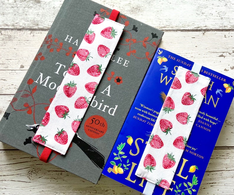 May include: Two book marks with a white background and red strawberries printed on them. The bookmarks are on top of two books. The book on the bottom is blue with the title "Still Life" and the book on top is gray with the title "To Kill a Mockingbird 50th Anniversary Edition".