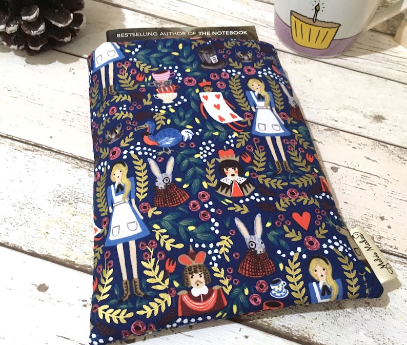 May include: A rectangular fabric book sleeve featuring a dark blue background with a repeating pattern of characters and foliage. The print includes figures in blue dresses, rabbits, playing cards, and crowns, with a small tag labelled Melis Makes.