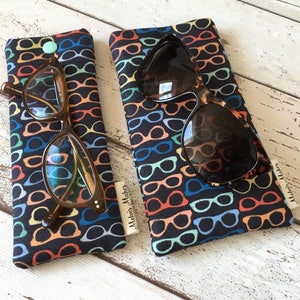 May include: Two glasses cases with a black background and a colorful pattern of eyeglasses. The cases are made of fabric and have a small tag that says "Maddie's Makers".