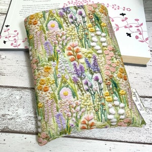 Puede incluir: Una funda de libro blanca con un dise&ntilde;o de bordado floral. El bordado presenta una variedad de flores en tonos de rosa, amarillo, naranja y morado. La funda del libro tiene una etiqueta que dice "Book Buddy".