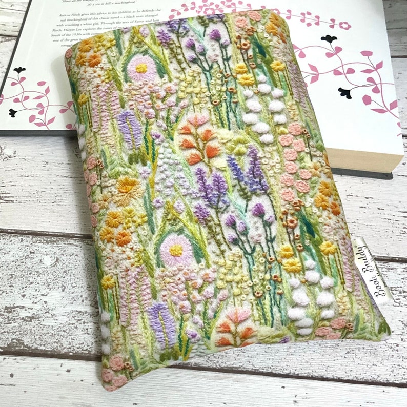 Puede incluir: Una funda de libro blanca con un dise&ntilde;o de bordado floral. El bordado presenta una variedad de flores en tonos de rosa, amarillo, naranja y morado. La funda del libro tiene una etiqueta que dice "Book Buddy".