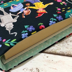 May include: A book sleeve featuring a colorful illustration of characters from The Wizard of Oz on a black background. The design includes Dorothy, the Tin Man, and the Scarecrow, with floral and tree accents. The interior is light green with white polka dots.