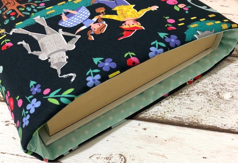 May include: A book sleeve featuring a colorful illustration of characters from The Wizard of Oz on a black background. The design includes Dorothy, the Tin Man, and the Scarecrow, with floral and tree accents. The interior is light green with white polka dots.