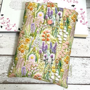 Puede incluir: Una funda de libro blanca con un dise&ntilde;o de bordado floral. El bordado presenta una variedad de flores en tonos de rosa, morado, amarillo y naranja. La funda de libro tiene un cierre de bot&oacute;n rosa y una etiqueta que dice "Book Buddy".