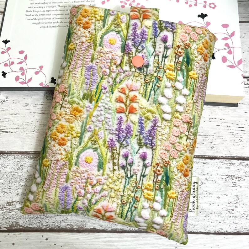 Puede incluir: Una funda de libro blanca con un dise&ntilde;o de bordado floral. El bordado presenta una variedad de flores en tonos de rosa, morado, amarillo y naranja. La funda de libro tiene un cierre de bot&oacute;n rosa y una etiqueta que dice "Book Buddy".