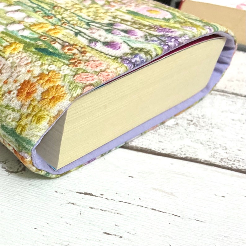 Puede incluir: Una cubierta de libro con un patr&oacute;n floral en tonos de verde, amarillo, rosa y morado. La cubierta est&aacute; hecha de tela y tiene un borde morado claro. El libro est&aacute; cerrado y la columna vertebral es visible.