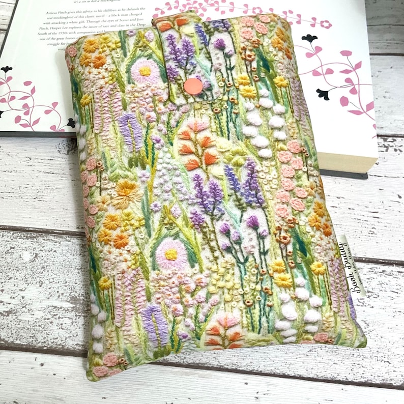 Puede incluir: Una funda de tel&eacute;fono de tela blanca con un dise&ntilde;o de bordado floral. La funda presenta una variedad de flores en tonos de rosa, morado, amarillo y naranja. La funda tiene un peque&ntilde;o bot&oacute;n rosa en la parte delantera.