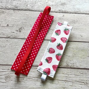 May include: Two fabric book markers. One is red with white polka dots and a red elastic band. The other is white with a strawberry print and a white elastic band.