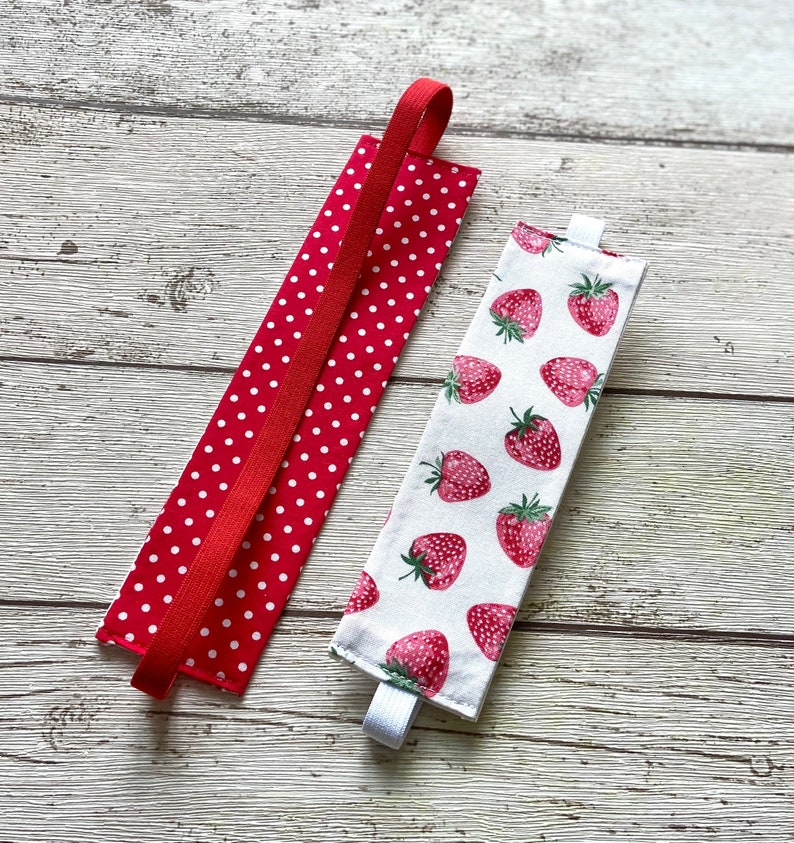 May include: Two fabric book markers. One is red with white polka dots and a red elastic band. The other is white with a strawberry print and a white elastic band.