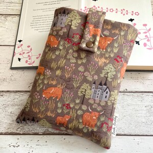 Può includere: Una custodia per libri in tessuto marrone con un motivo di mucche delle Highlands e castelli. La custodia ha una chiusura a bottone ed è appoggiata su un libro aperto. Il design include mucche arancioni, castelli grigi e fiori rossi.