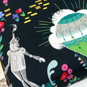 May include: A fabric with a whimsical design featuring the Tin Man from The Wizard of Oz. The illustration includes colorful flowers, a hot air balloon, and a dark background. The Tin Man is depicted in a silver color.