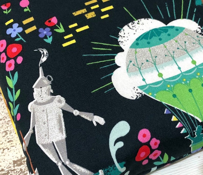 May include: A fabric with a whimsical design featuring the Tin Man from The Wizard of Oz. The illustration includes colorful flowers, a hot air balloon, and a dark background. The Tin Man is depicted in a silver color.