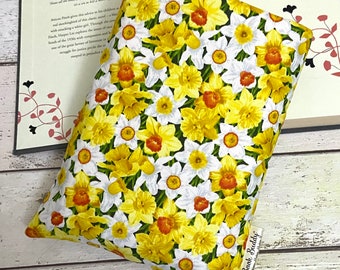 以下が含まれることがあります： 黄色と白の水仙の花柄が鮮やかな長方形のブックカバー。布製のカバーが開いた本の上に置かれています。「Book Buddy」の文字が入った小さなタグが付いています。