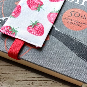 May include: A red and white fabric bookmark with a strawberry pattern. The bookmark is attached to a gray book with the text "50th Anniversary Edition" on the cover.