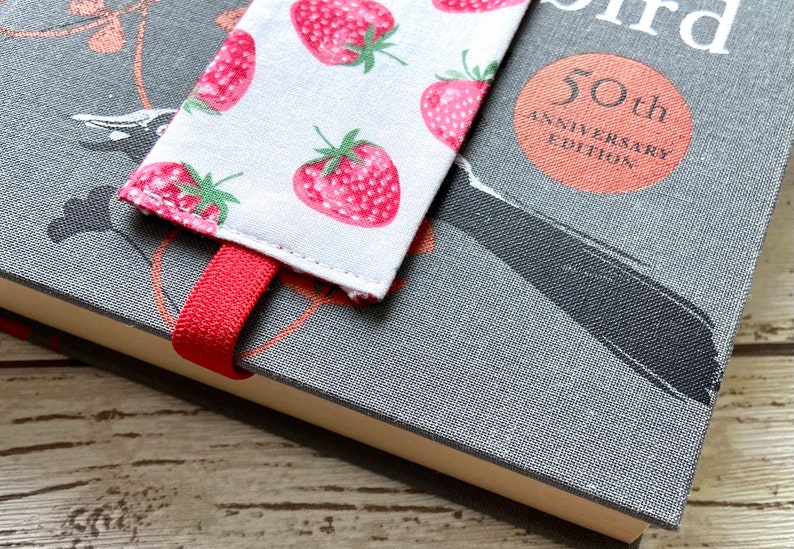 May include: A red and white fabric bookmark with a strawberry pattern. The bookmark is attached to a gray book with the text "50th Anniversary Edition" on the cover.