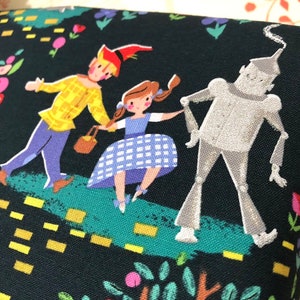 May include: A black fabric featuring a colorful illustration of characters from The Wizard of Oz. The illustration includes Dorothy in a blue gingham dress, the Scarecrow, and the Tin Man, set against a whimsical background of flowers and a yellow brick road.