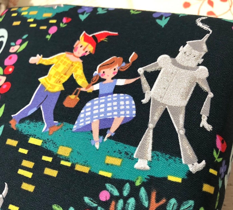 May include: A black fabric featuring a colorful illustration of characters from The Wizard of Oz. The illustration includes Dorothy in a blue gingham dress, the Scarecrow, and the Tin Man, set against a whimsical background of flowers and a yellow brick road.