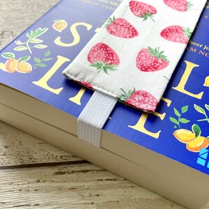 May include: A blue hardcover book with the title "Small Life" in yellow lettering. The book has a white elastic bookmark with a fabric bookmark featuring a strawberry pattern. The book is on a wooden surface.