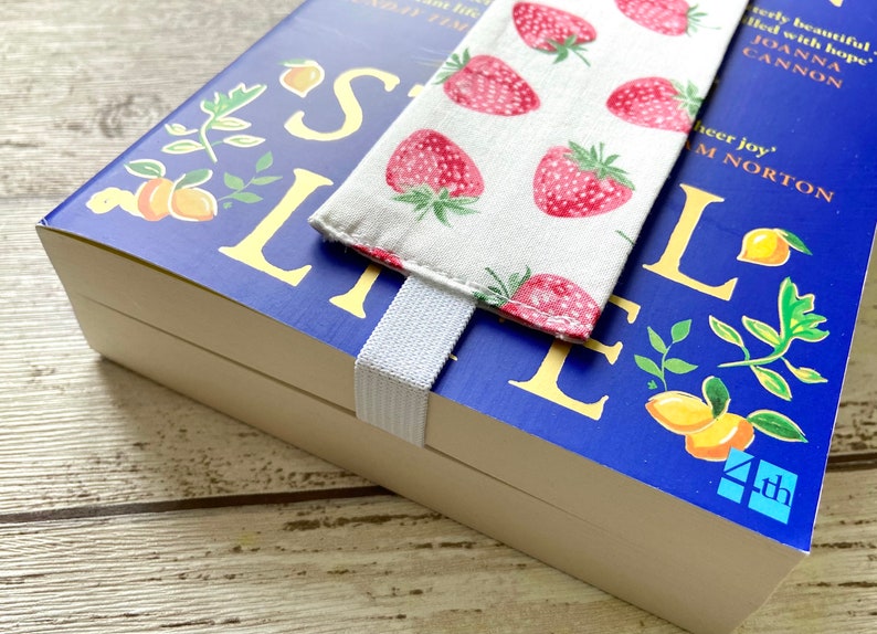 May include: A blue hardcover book with the title "Small Life" in yellow lettering. The book has a white elastic bookmark with a fabric bookmark featuring a strawberry pattern. The book is on a wooden surface.
