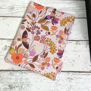Puede incluir: Una funda de libro de tela beige con un patrón floral en tonos naranja, rosa y marrón. La funda presenta un pequeño diseño de búho y un cierre de botón morado. El texto "Melina Makes" está impreso en una etiqueta rosa.